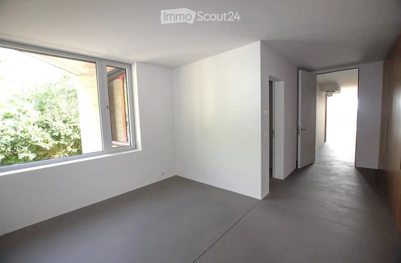Empty room with white walls, grey tiled floor, large window with view of greenery, and open doorway leading to another space.