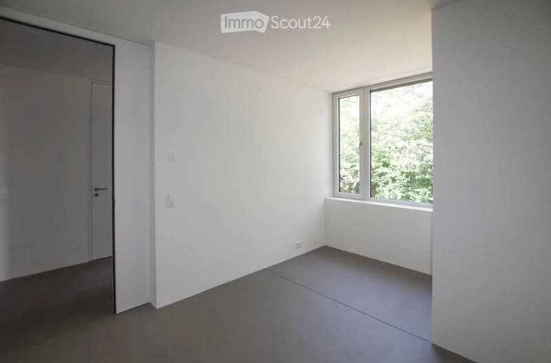 Empty room with white walls, grey floor, and a window with a view of greenery.