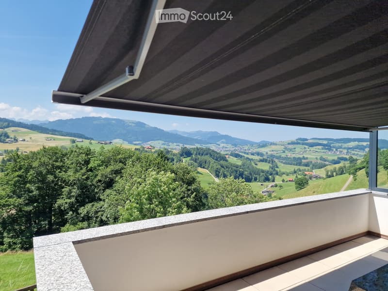 Balcony with a panoramic view of green fields, trees, and distant mountains under a clear blue sky.