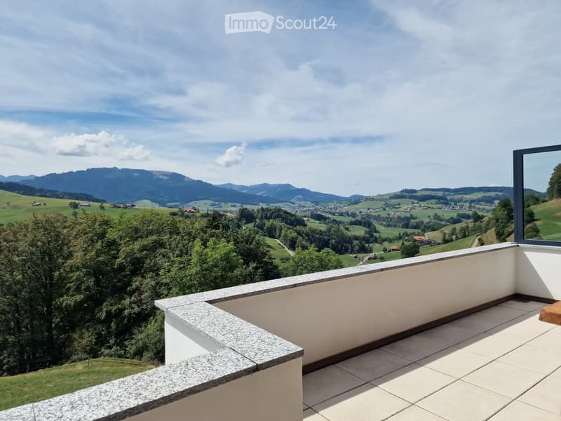 Balcony with tiled floor and stone railing, offering a scenic view of green fields, trees, and distant mountains under a partly cloudy sky.