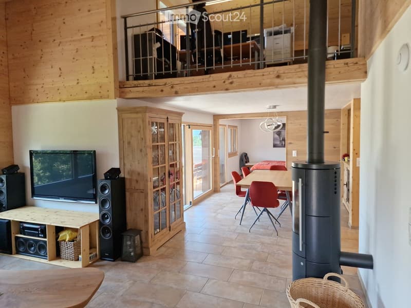A spacious, open-concept living and dining area with wooden walls, ceiling, and flooring. Features include a large TV with speakers, a wooden cabinet, a dining table with red chairs, and a modern wood-burning stove. The area is well-lit with natural light