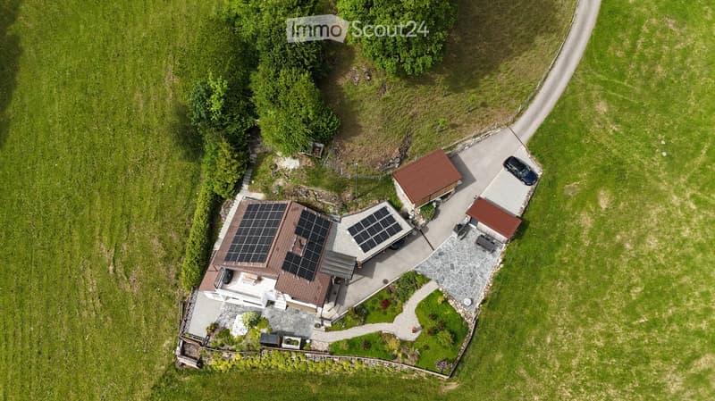 Aerial view of a modern house with solar panels on the roof, surrounded by a well-maintained garden and driveway. Includes a small outbuilding and a parked car.