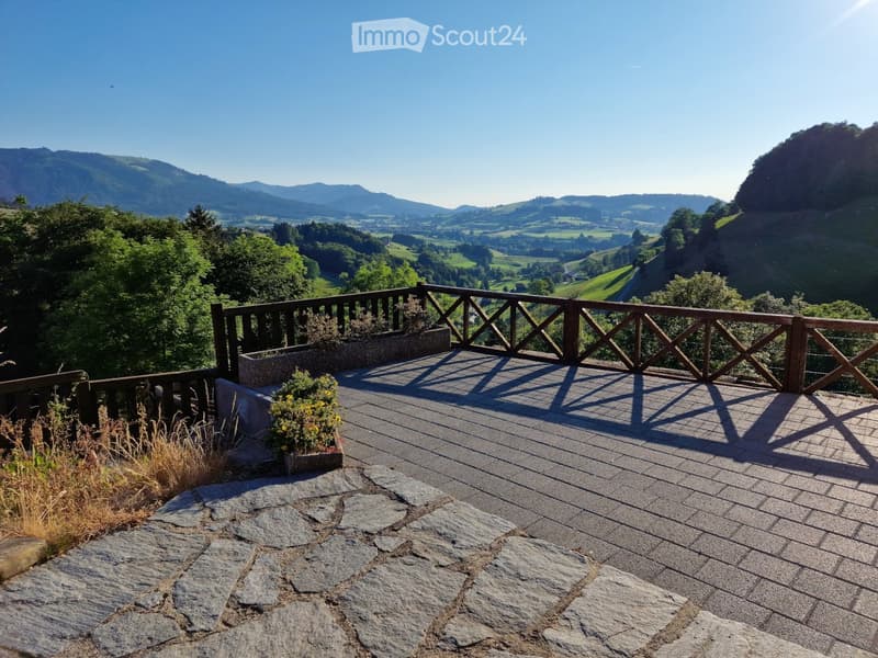Wooden deck with railing, overlooking a scenic valley with mountains and greenery.