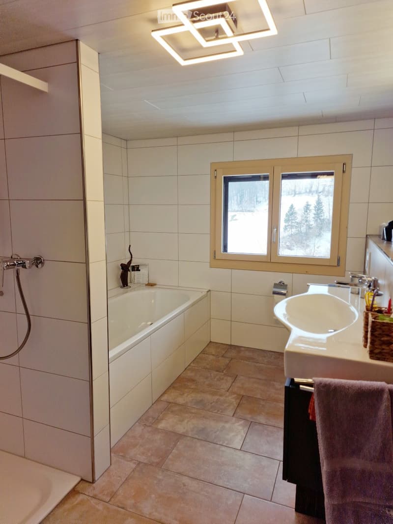 Modern bathroom with a bathtub, sink, and large window overlooking a snowy landscape. Features tiled walls and floor, a shower area, and a towel on a rack.