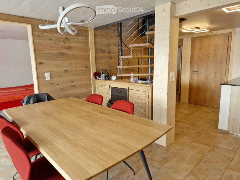 Modern, open-plan living and dining area with wooden walls, a large wooden dining table with red chairs, a red sofa, and a small cabinet with various items on top.
