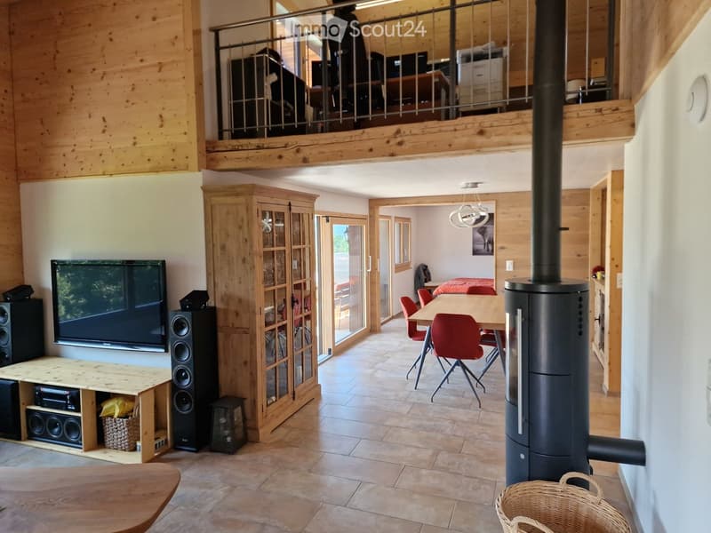 Open-plan living and dining area with wooden beams, TV, speakers, dining table with chairs, and a modern wood-burning stove.