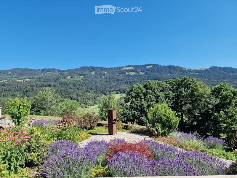 A well-maintained garden with a variety of colorful flowers, lavender bushes, and a metal sculpture, set against a backdrop of rolling hills and dense forest.