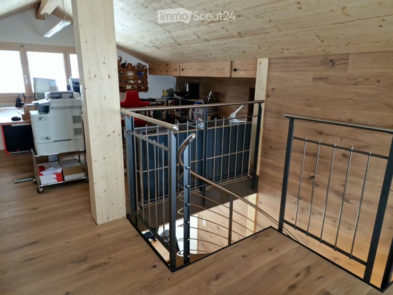 Open-plan office space with wooden flooring, exposed wooden beams, a modern staircase with metal railing, and a desk with office equipment.