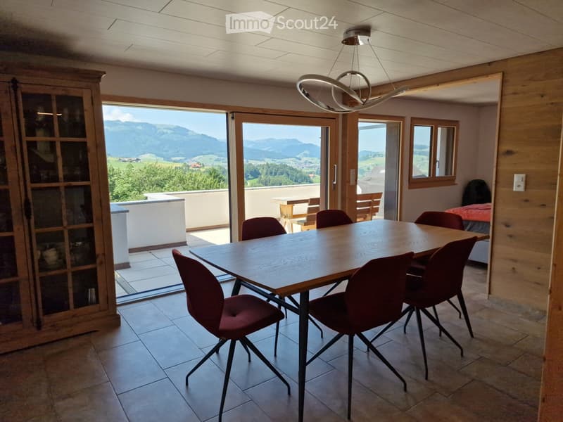 Bright dining area with a wooden table and red chairs, large windows offering a scenic mountain view, and a modern pendant light fixture.