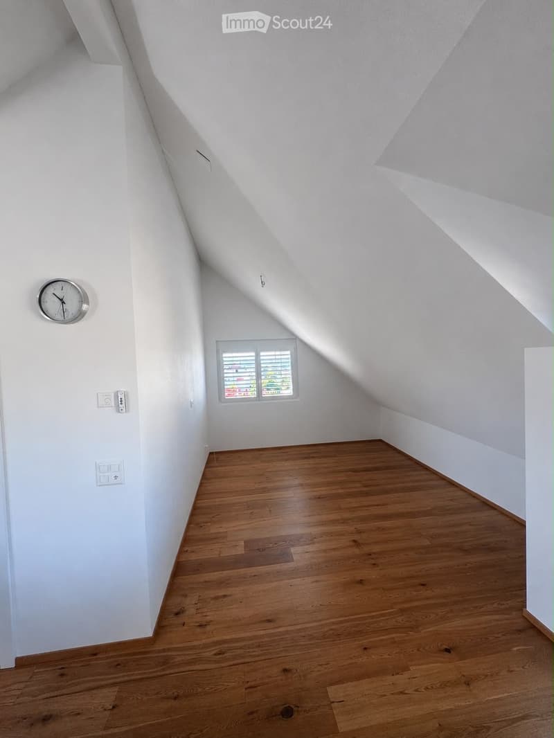Empty attic room with wooden flooring, white walls, and a small window with blinds. The ceiling has a sloped design typical of attics.