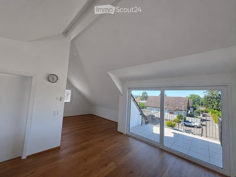 Bright, empty attic room with wooden flooring, white walls, and a large sliding glass door leading to a balcony. The room features a slanted ceiling and a small window on the left side.