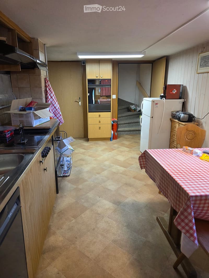 A small kitchen with tiled flooring, wooden cabinets, a refrigerator, a table with a checkered tablecloth, and a chair. The kitchen features a sink, stove, and microwave.