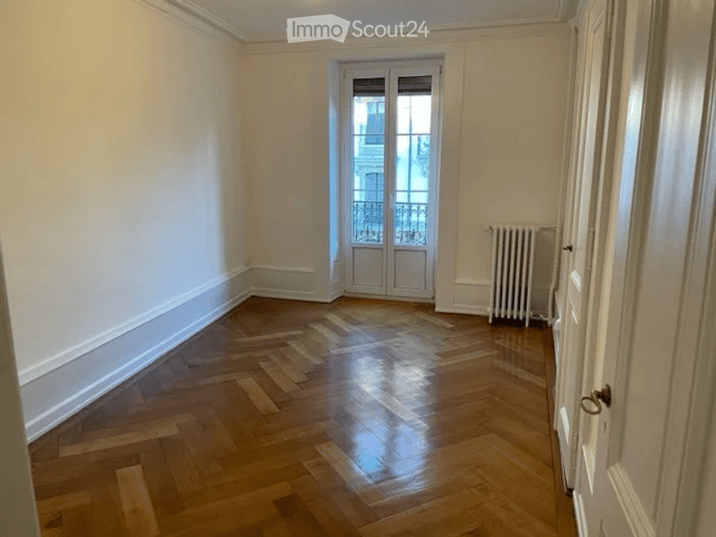 Empty room with wooden parquet flooring, white walls, a radiator, and a window with shutters.