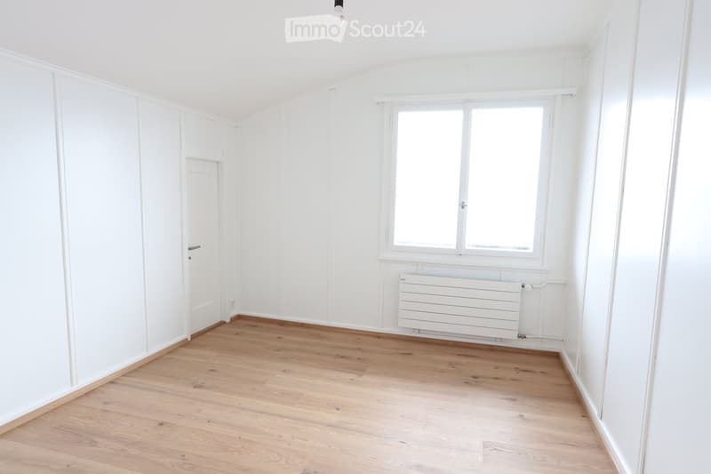 Empty room with white walls, wooden floor, a window, a door, and a radiator.