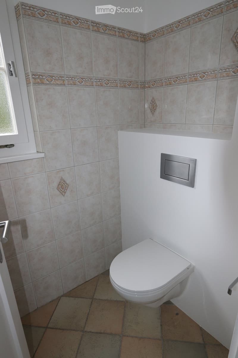 Modern bathroom with white toilet, beige tiled walls, and decorative border tiles. Features a window with frosted glass and a chrome flush plate.