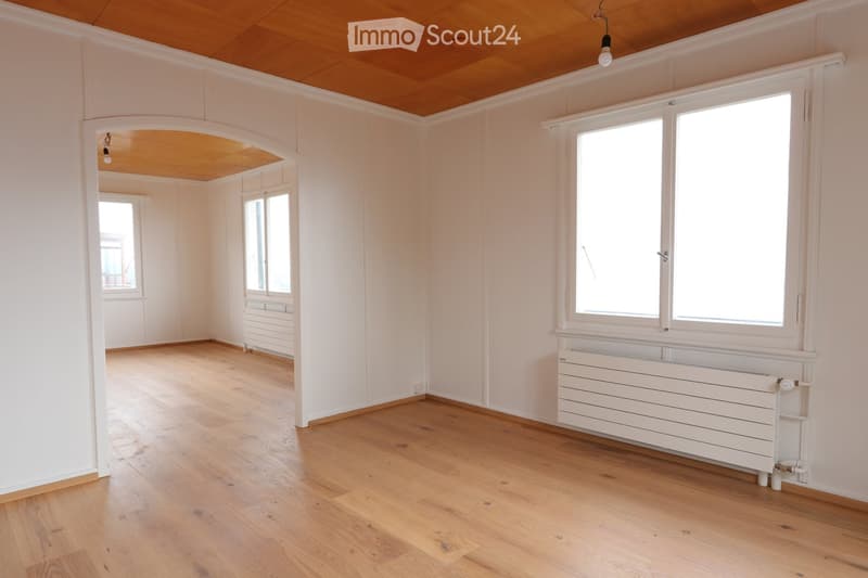 Empty room with light wooden flooring, white walls, large windows, and a white radiator. The room has a wooden ceiling and an arched doorway leading to another space.
