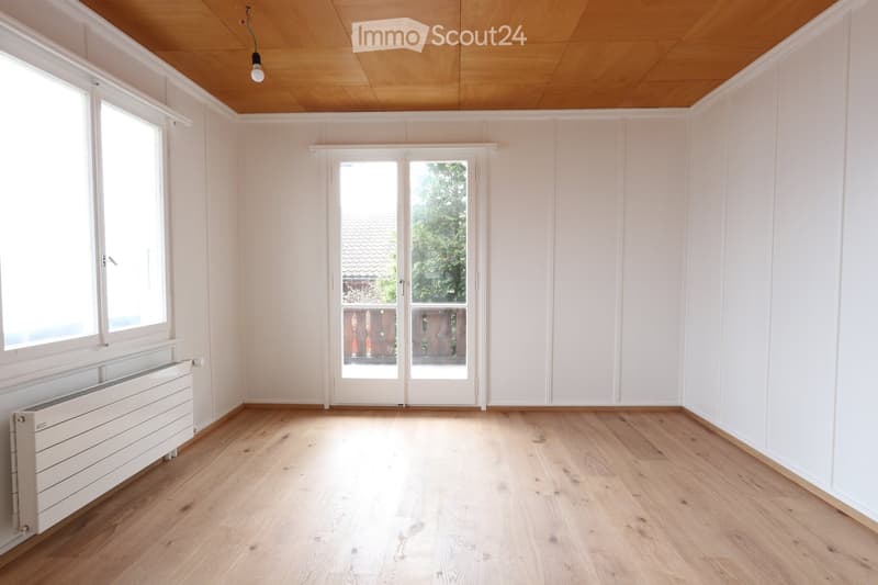 Empty room with light wooden flooring, white walls, large windows, and a sliding glass door leading outside.