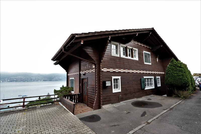 A two-story wooden house with white-framed windows, green shutters, and a view of a body of water in the background.