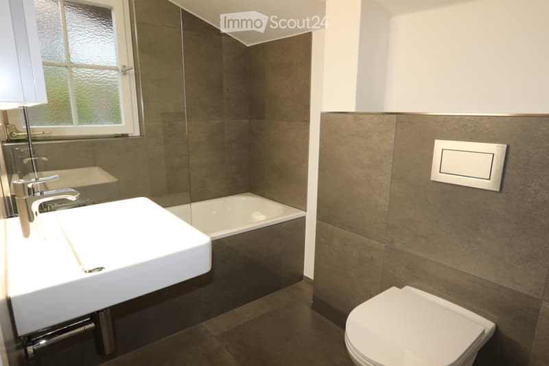 Modern bathroom with a white sink, chrome faucet, bathtub, and toilet. Features grey tiled walls and a frosted window.