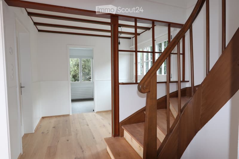 Bright hallway with wooden flooring, white walls, and a wooden staircase leading upwards. Features include a glass-paneled door at the end and a light fixture hanging from the ceiling.