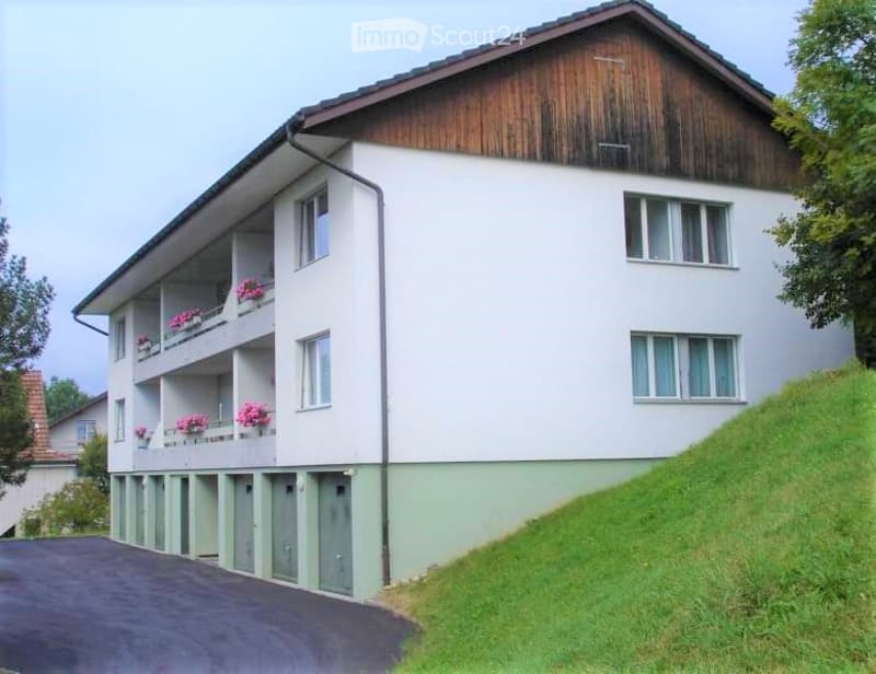 Two-story building with white walls, wooden roof, multiple balconies with flower boxes, and a paved driveway.