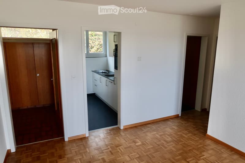 Bright hallway with wooden flooring, white walls, and several doors leading to different rooms, including a kitchen area visible in the background.