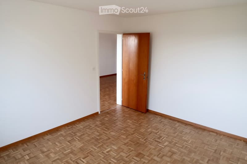 Empty room with light-colored walls and wooden herringbone flooring. A wooden door is open, revealing another room with similar flooring.