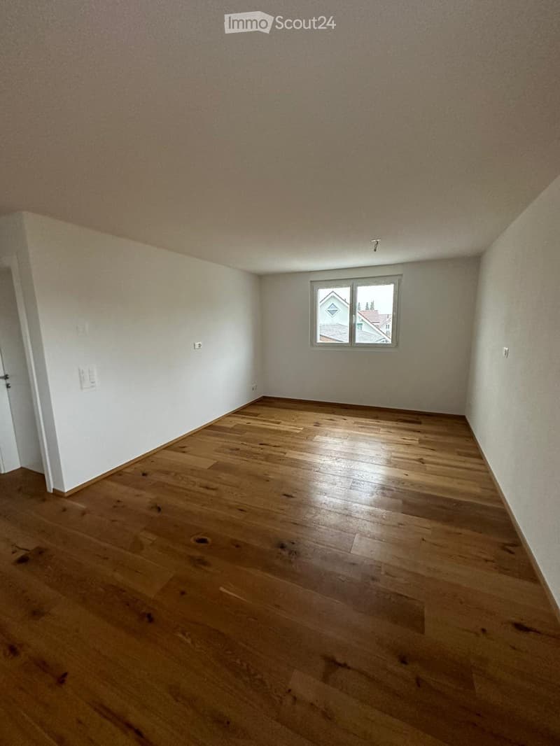 Empty room with wooden flooring, white walls, and a single window showing a view of neighboring buildings.