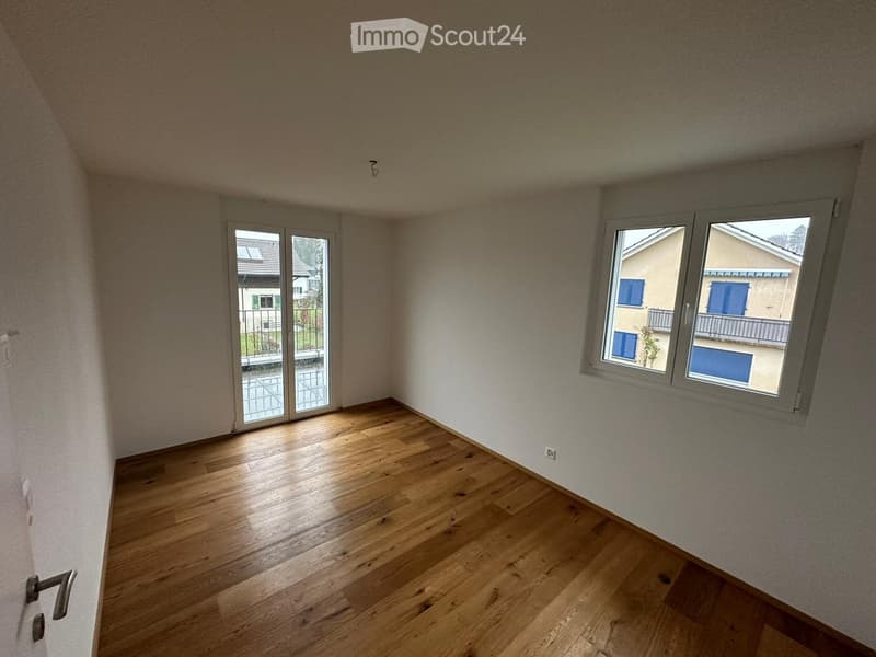 Empty room with wooden flooring, large windows, and a sliding glass door leading to a balcony.