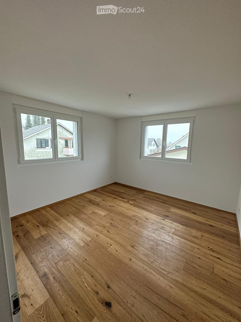 Empty room with wooden flooring and white walls, featuring two windows with grey frames overlooking neighboring houses.
