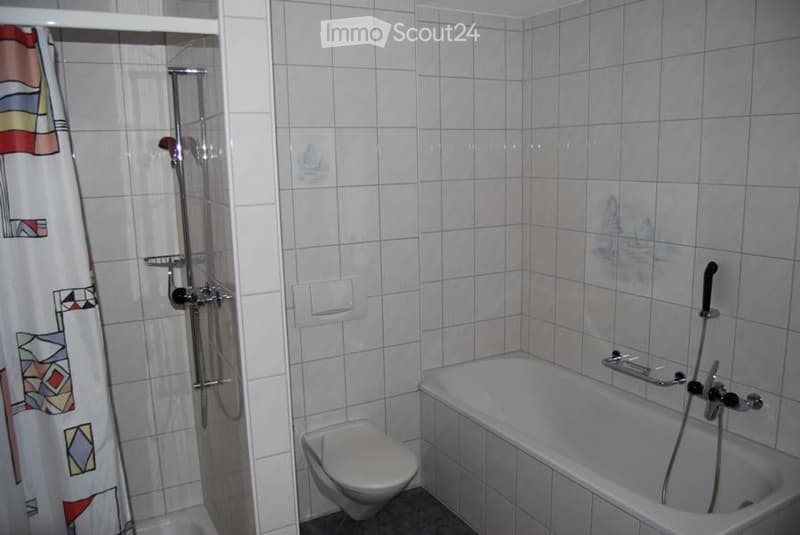 Bathroom with white tiled walls and floor, featuring a bathtub with a handheld shower, a toilet, and a shower curtain with a colorful pattern.