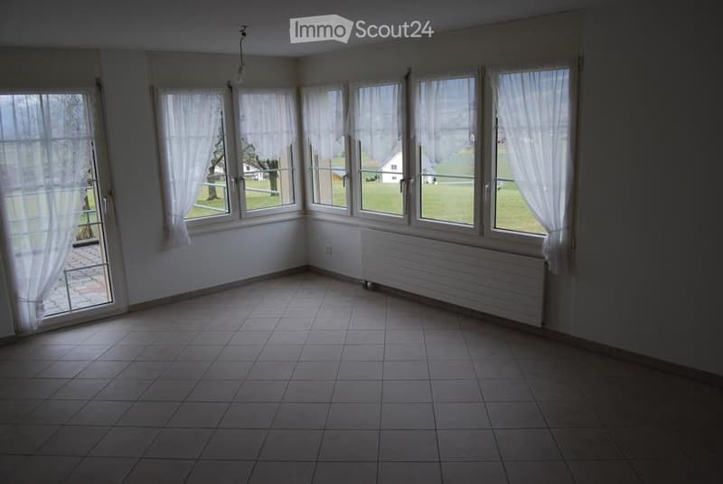 Empty room with tiled floor, large windows with white curtains, radiator, and a view of a green landscape.