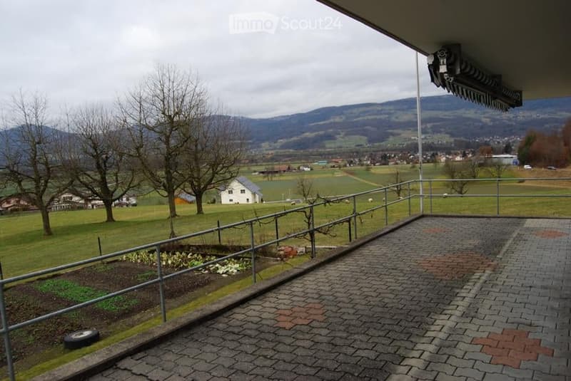 Balcony with a view of a rural landscape, including fields, trees, and distant mountains. The balcony is paved with bricks and has a metal railing.