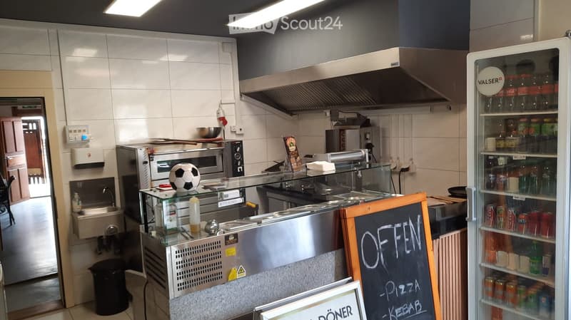 Modern kitchen with stainless steel appliances, a refrigerator, an oven, a sink, and a menu board displaying 'OFFEN - PIZZA - KEBAB'.