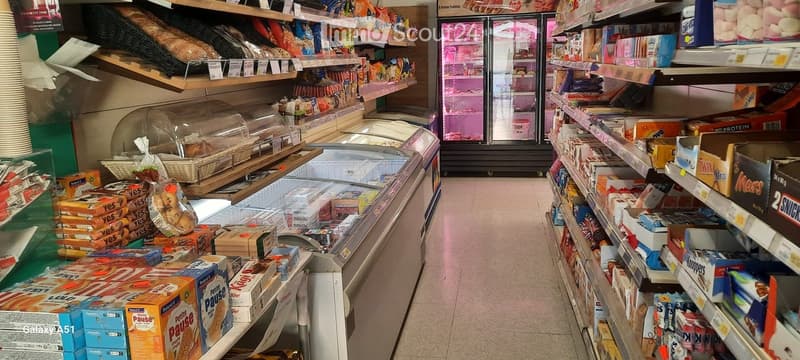 A small convenience store interior with shelves stocked with snacks, drinks, and bread, and a refrigerator unit in the background.