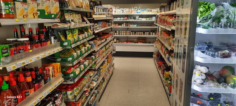 A grocery store aisle with shelves stocked with various food products, including sauces, condiments, and packaged goods. A refrigerator unit is visible on the right side with fresh produce inside.