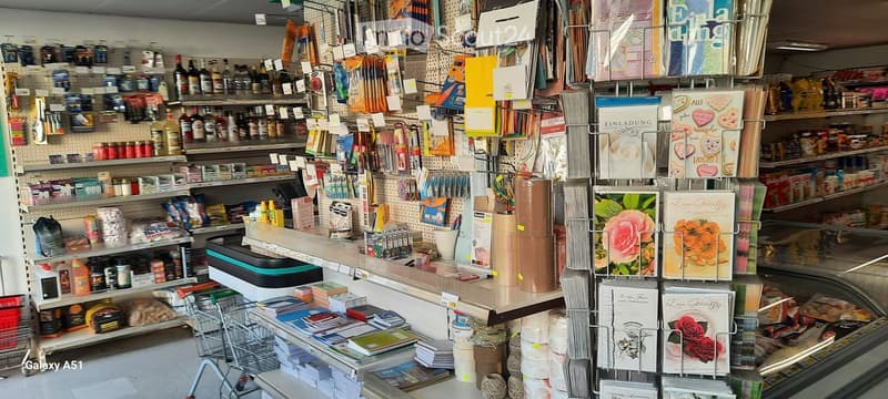 A well-stocked stationery and gift shop with shelves filled with various products, including stationery, greeting cards, and household items. A display stand with greeting cards is visible, along with a cash register and a shopping cart.