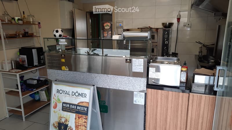 Commercial kitchen with stainless steel appliances, including a refrigerator, oven, microwave, and counter. Shelves hold various items, and a signboard advertises 'Royal Döner'.