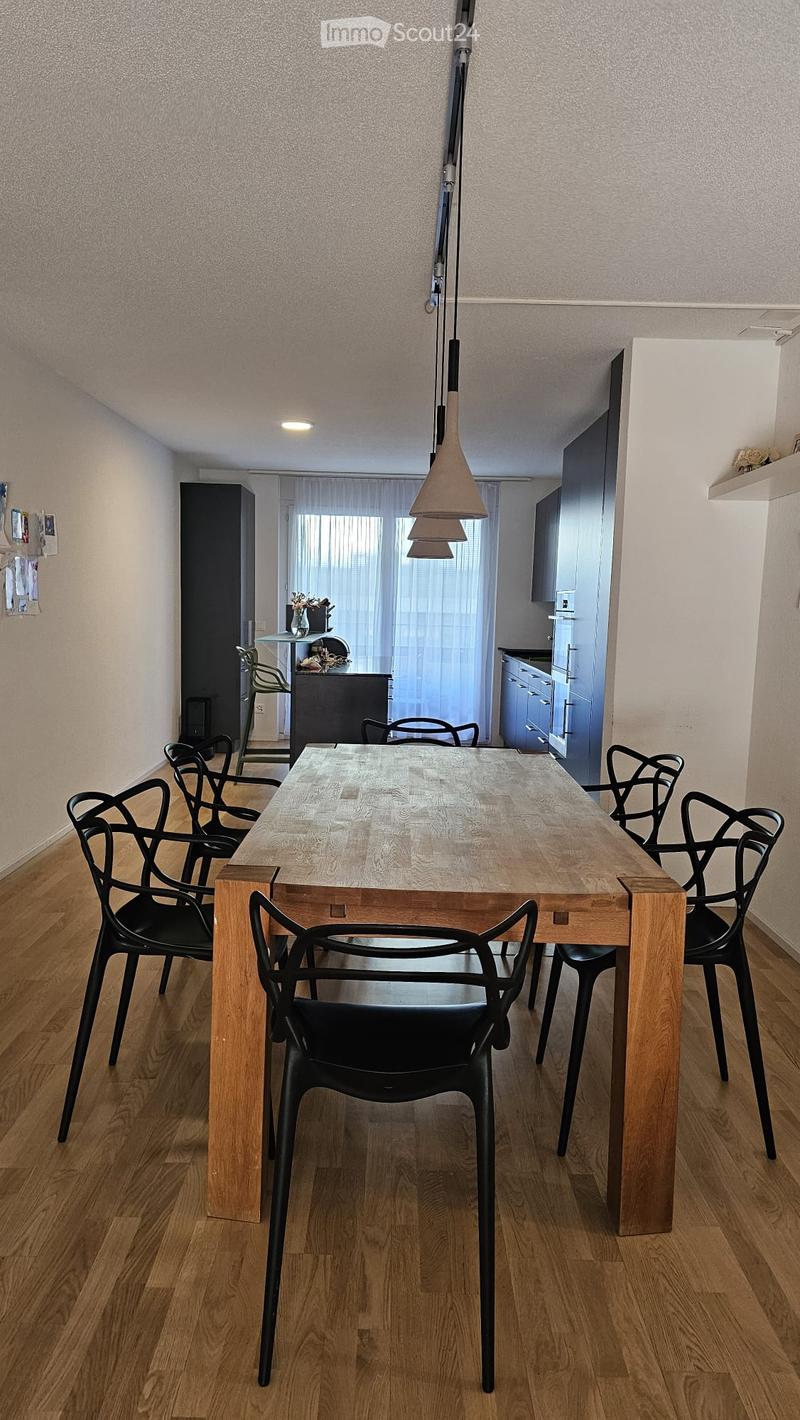 Dining area with a wooden dining table surrounded by black chairs, a hanging pendant light, and a view into a kitchen area with cabinets and a window with curtains.