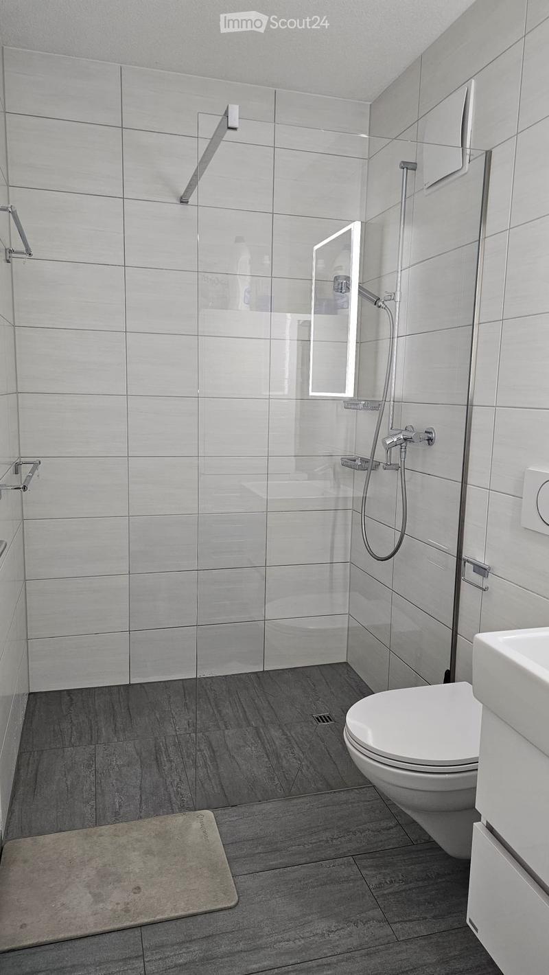Modern bathroom with white tiles, a glass-enclosed shower, a toilet, a sink, and a bathroom rug on the floor.