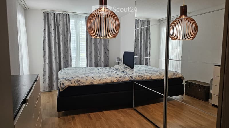 Modern bedroom with a large bed, nightstand, mirror, wardrobe, and pendant lighting.