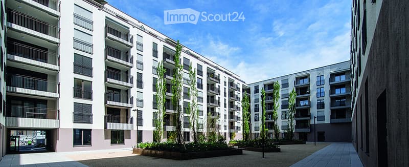 Modern multi-story residential building with balconies and contemporary design, surrounded by greenery and paved walkways.