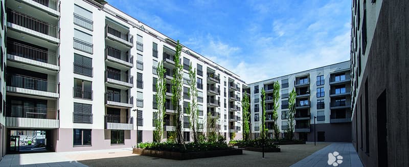 Modern multi-story residential building with balconies and contemporary design, surrounded by greenery and paved walkways.