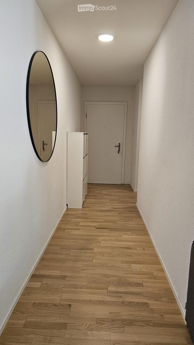 A narrow hallway with wooden flooring, white walls, and a single round mirror on the left side. A closed door is visible at the end of the hallway.