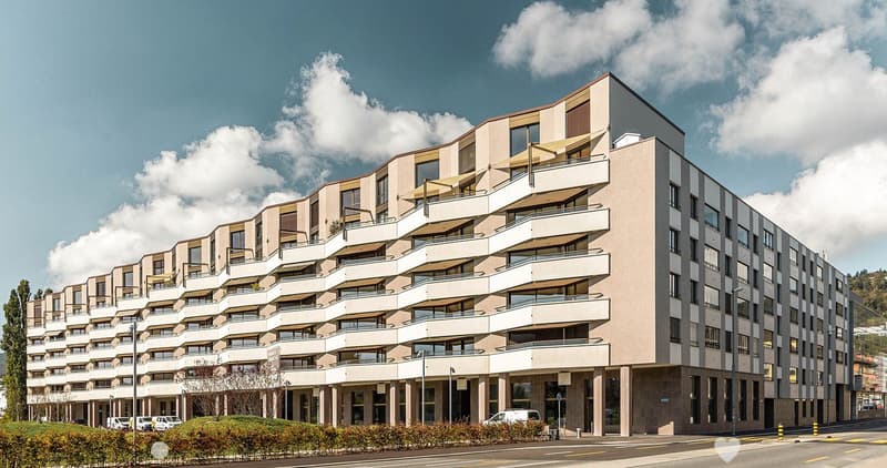 Modern multi-story residential building with balconies and large windows, featuring a contemporary design with beige and grey tones.