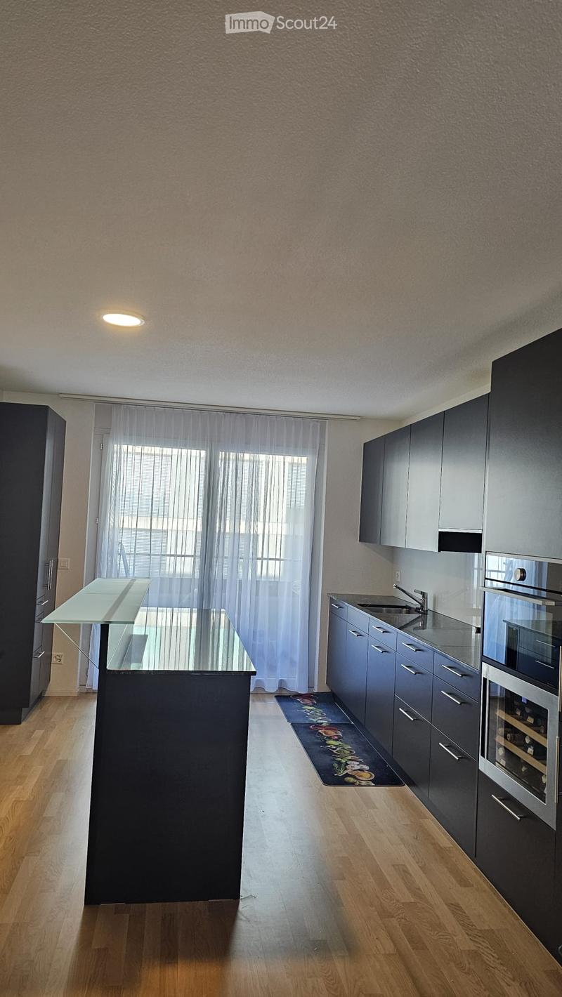 Modern kitchen with dark grey cabinets, a central island with a glass top, a sink with a faucet, and a wine cooler. The floor is wooden, and there is a small rug near the sink area.