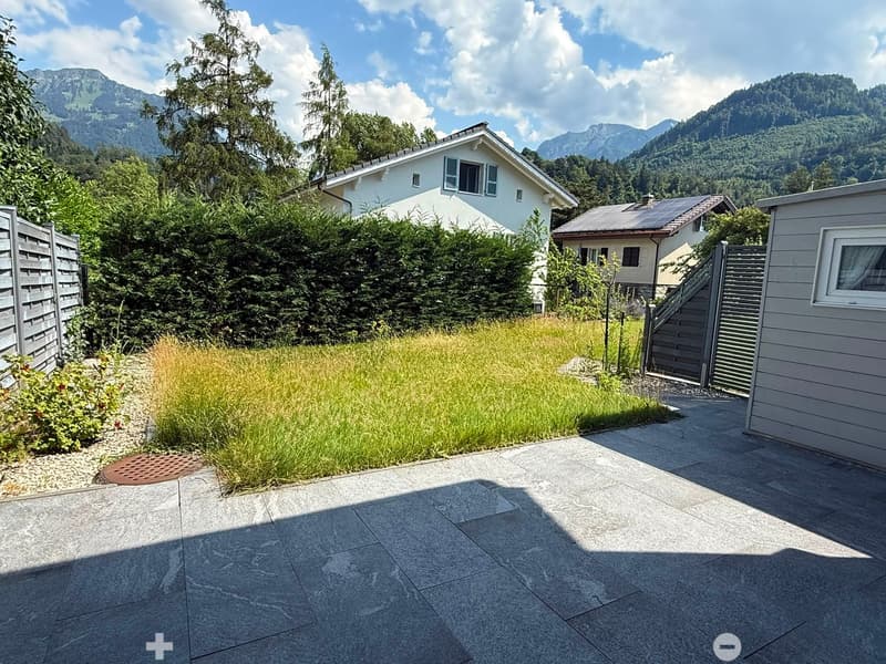 Garden area with a paved patio, grassy patch, small plants, and a wooden fence, with houses and mountains in the background.