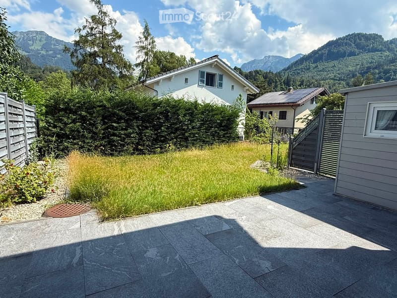 Garden area with a paved patio, grassy patch, small plants, and a wooden fence, with houses and mountains in the background.