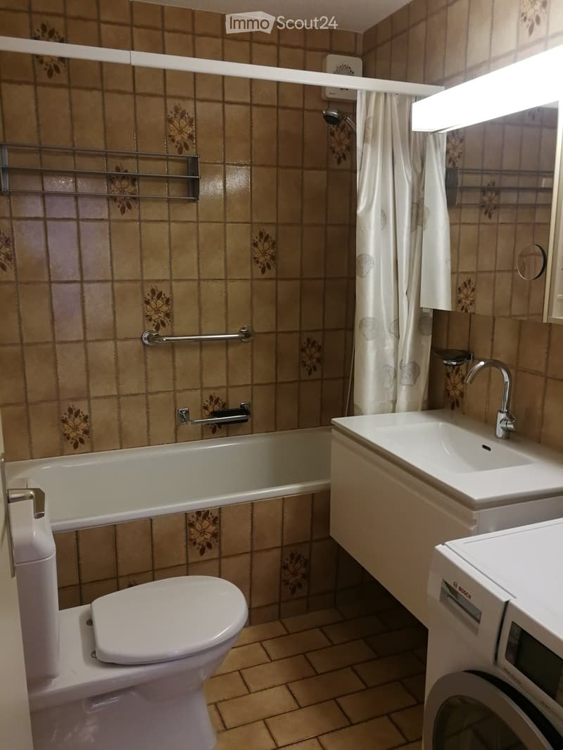 Bathroom with beige tiled walls and floor, featuring a toilet, bathtub, sink, and washing machine. Includes a shower area with a curtain and a metal rack.