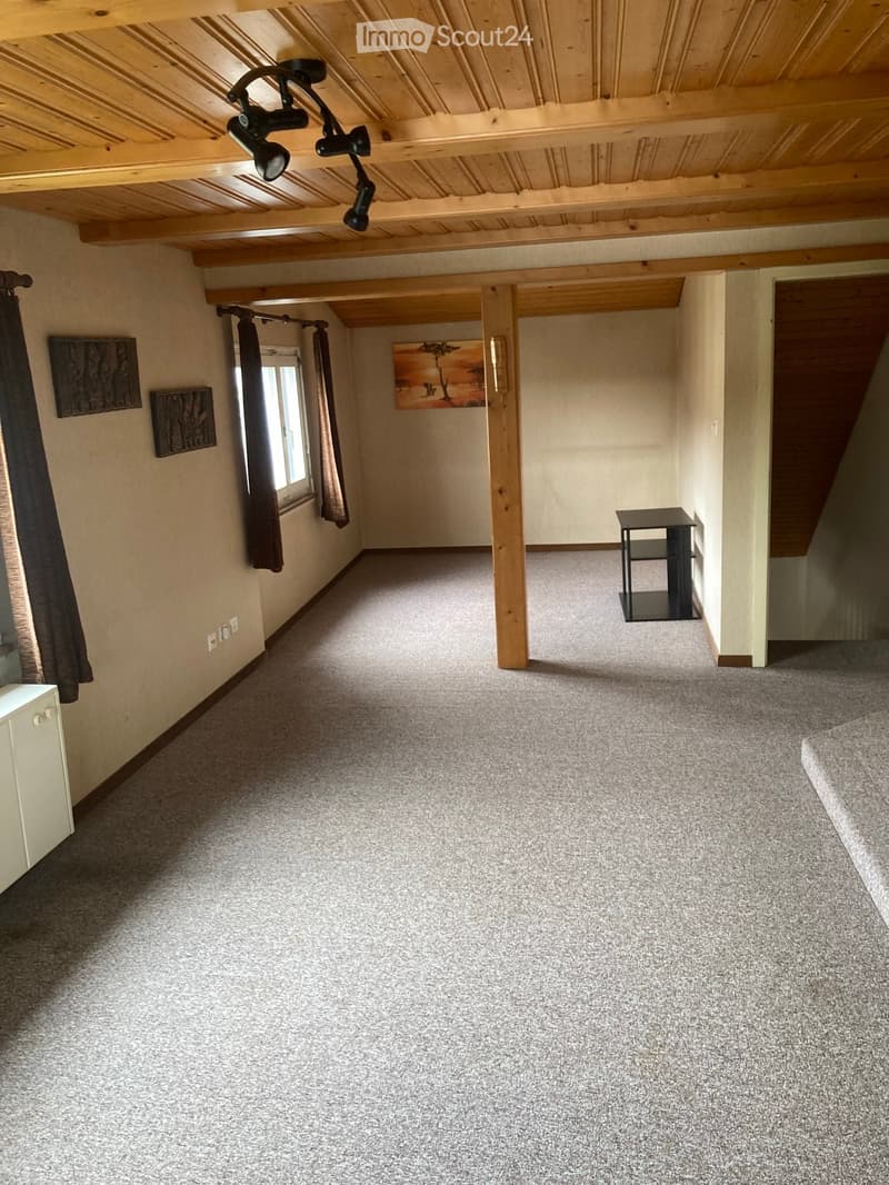 Empty room with carpeted floor, wooden beams, exposed ceiling lights, and a small black table near the wall.